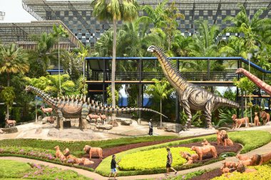 Chon Buri, Tayland - 5 Eylül 2023: Nong Nooch Tropik Botanik Bahçesi 'ndeki Dinozor Vadisi' ndeki çeşitli boyutlardaki güzel dinozor heykellerinde turistler, başka bir büyük turistik ilgi merkezi..