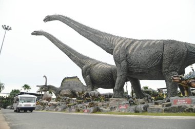 CHON BURI, THAILAND - 6 Eylül 2023: Nong Nooch Tropikal Bahçesi 'ndeki Dinozor Vadisi' ndeki çeşitli boyutlardaki güzel dinozor heykellerinde turist otobüsü, başka bir büyük turistik ilgi alanı..