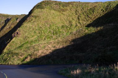 Yeşil bir yamaç güneş ışığıyla yıkanmış, gölge yamaları çarpıcı bir kontrastlık yaratıyor. Çimenli arazi, ışığın ve gölgenin etkileşimiyle vurgulanır, manzaranın doğal güzelliği.