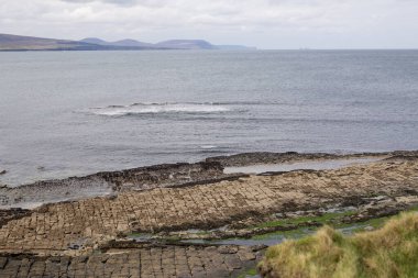 Bulutlu bir gökyüzünün altında çarpıcı kayalık bir kıyı şeridi ve sakin sular barındıran pitoresk bir kıyı manzarası.