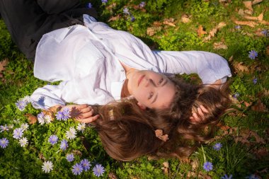Çiçeklerle çevrili yemyeşil çimlerin üzerinde mutlu ve huzurlu bir şekilde dinlenen bir kadının sahnesi.