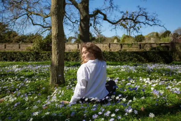 Bir kadın, berrak mavi gökyüzünün altında canlı bahar çiçekleriyle çevrili yemyeşil çimlerin üzerinde zarifçe oturuyor.