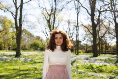 Genç bir kadın zarif bir manzarada duruyor, güzel çiçekler ve uzun ağaçlarla çevrili.