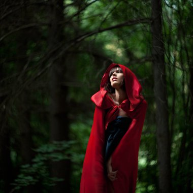 Çarpıcı kırmızı pelerinli bir kadın büyüleyici bir ormanda zarif bir şekilde duruyor ve büyüleyici bir sahne yaratıyor.