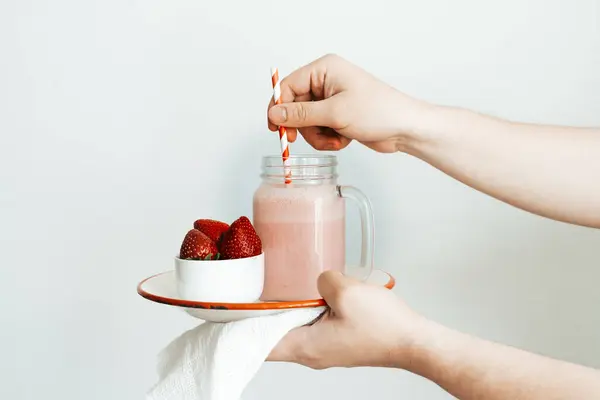 Beyaz arkaplanda bir kavanoz çilekli smoothie ile tabak tutan bir el.