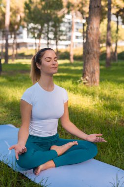 Yoga minderinde oturmuş parkta meditasyon yapan genç bir kadın.