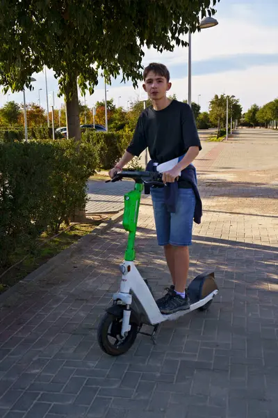 Asfalt yolda elektrikli scooter kullanan genç bir adam. O, siyah uzun kollu bir gömlek ve sıradan bir duruşu olan kot şort giyiyor. Arkaplanda ağaçlar ve açık bir gökyüzü var.