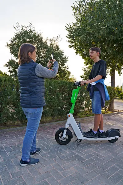 Parkta elektrikli scooterlı bir çocuğun fotoğrafını çeken bir kadın. Kadın anı yakalamak için akıllı telefon tutarken, çocuk scooter 'ın üzerinde günlük giysiler giyiyor. Yeşil ağaçlar