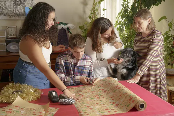 Bir aile hediye paketlemek için masanın etrafında toplanır. Bir kız bakarken, iki kadın ve bir genç çocuk ambalaj kağıdı kesmekle meşguller. Kadınlardan birinin yanında bir köpek oturuyor ve şenlik süsleri...