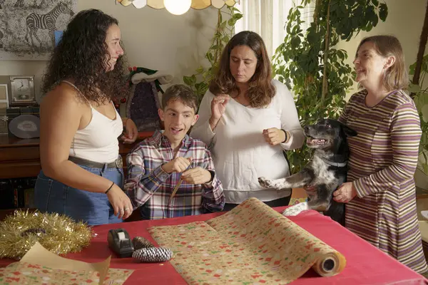 Dört kişinin hediye paketlediği bir aile toplantısı. İki kadın ve bir çocuk paketleme yaparken, bir köpek neşe içinde etkileşim halindedir. Dekorasyon ve sıcak bir atmosferle şenlik havası var.