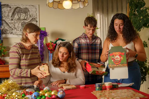 Tatil için dekore edilmiş bir masanın etrafında toplanan bir aile. Dört kişi renkli ambalaj kağıtları ve süslemelerle hediyeleri paketlemekle meşgul. Atmosfer şenlikli ve neşeli.