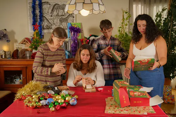 Tatil için dekore edilmiş bir masanın etrafında toplanan bir aile hediye paketleme faaliyetleriyle meşgul. Renkli süsler, ambalaj kağıtları ve masaya saçılmış şenlikli dekorasyonlar var. ATM 'de.