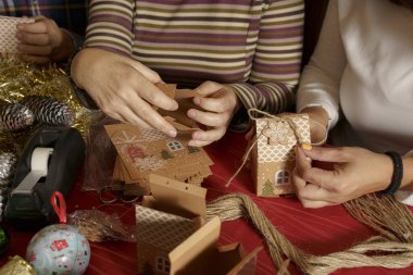 İki kişi bayram hediyesi kutuları hazırlıyor. Şenlik tasarımlarıyla süslenmiş. Etrafları kurdele, bant ve süs gibi el yapımı malzemelerle çevrili. Sıcak bir tatil ortamı yaratıyorlar..