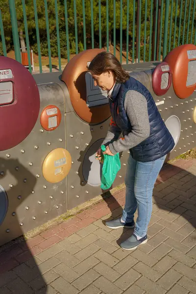 Bir kadın bozuk para makinesini temizliyor. Makinenin üzerinde yeşil bir çanta var. Kadın mavi bir ceket giyiyor.