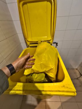 Bir kişi sarı bir çanta tutuyor ve onu sarı bir çöp kutusuna koyuyor. Çöp kutusu bir banyoda.