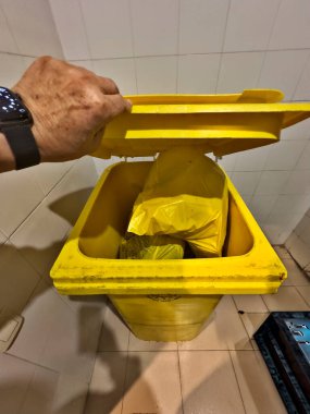 A person is holding a yellow trash can lid and pulling it open. The trash can is full of plastic bags clipart