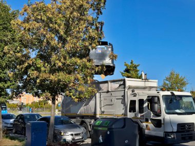 Ağaçlar ve açık mavi gökyüzü ile çevrili bir kamyonun arkasına araç çeken bir çekici. Sahne, park halindeki arabaların ve arka planda bir yerleşim alanının olduğu kentsel yaşamı gösteriyor..