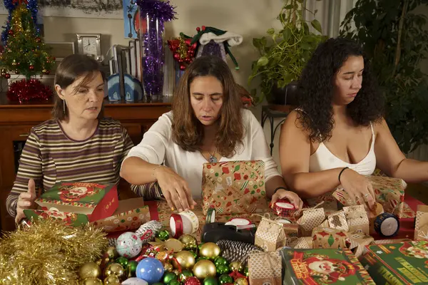 Bayram şenliği sırasında hediye paketlemesi yapan üç kadın. Etrafları renkli süslemeler, paketlenmiş hediyeler ve bir masanın üzerindeki çeşitli süslemelerle çevrili. Atmosfer neşeli ve sıcak.