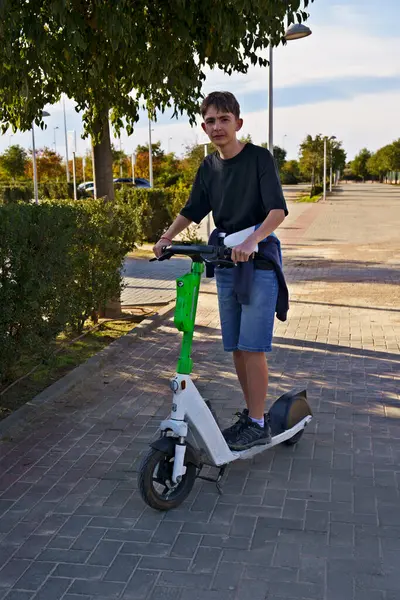 Parkta kaldırımlı bir yolda elektrikli scooter kullanan genç bir adam. O, beline bir kazak bağlanmış siyah bir gömlek ve kot şort giyiyor. Arka planda ağaçlar ve berrak bir gökyüzü var.