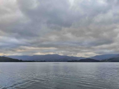 Bulutlu bir gökyüzünün altında dağlarla çevrili sakin bir göl manzarası. Su bulutlu gökyüzünü yansıtır ve sakin bir atmosfer yaratır..