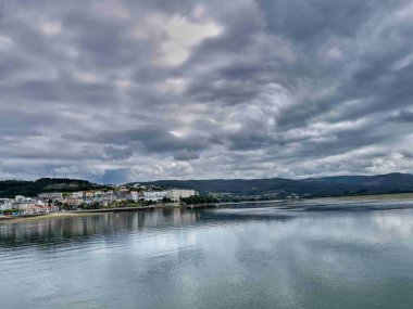 Sakin sulara yansıyan bulutlu gökyüzü ile kıyı kasabasının manzarası.