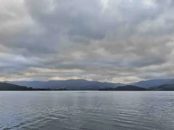 Bulutlu bir gökyüzünün altında dağlarla çevrili sakin bir göl manzarası. Su bulutlu gökyüzünü yansıtır ve sakin bir atmosfer yaratır..