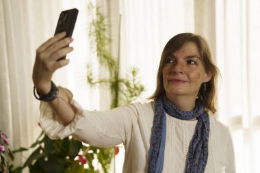 Bitkilerle dolu parlak bir odada akıllı telefonuyla selfie çeken bir kadın. Beyaz bir bluz ve mavi bir eşarp giyiyordu. Perdelerin arasından süzülen yumuşak doğal ışık vardı..