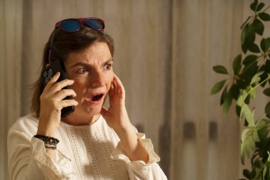 Akıllı telefondan konuşan şaşırmış bir kadın, eli yüzünde, arka planda bitkilerle aydınlık bir odada..