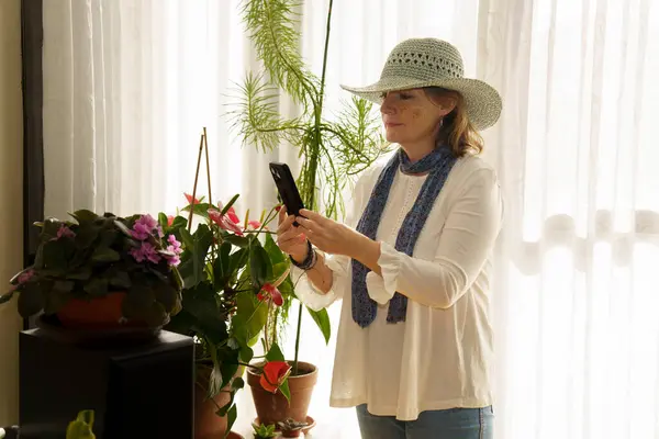 Geniş kenarlı şapka ve eşarp takan bir kadın elinde akıllı bir telefonla saksı bitkilerinin yanında duruyor. Güneş ışığı perdelerin arasından süzülüp sıcak bir atmosfer yaratır..