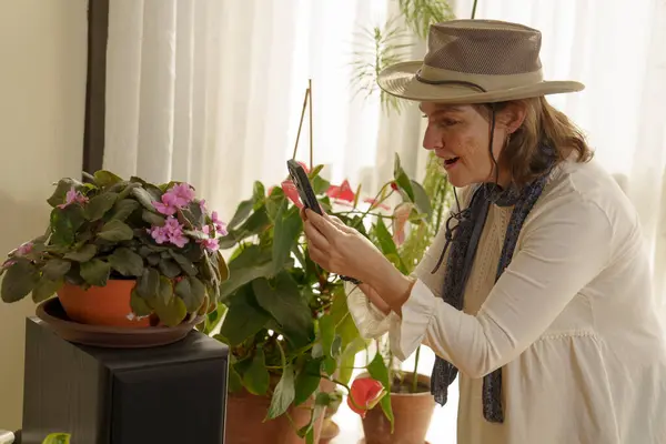 Şapkalı bir kadın evin içinde çiçeklerin açtığı, çeşitli ev bitkileriyle çevrili bir fotoğraf çekiyor. Güneş ışığı perdelerin arasından süzülür ve sıcak bir atmosfer yaratır..