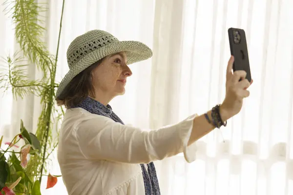 Geniş kenarlı şapka takan bir kadın akıllı telefonuyla selfie çeker. Kapalı alandaki bitkilerle çevrili, perdeleri olan bir pencerenin yanında duruyor. Işık yumuşak, sıcak bir atmosfer yaratır..