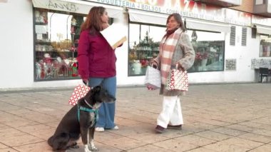 Alışveriş çantalı iki kadın, yanlarında siyah bir köpekle Madrid 'de bir kaldırımda alışverişlerini tartışıyor.