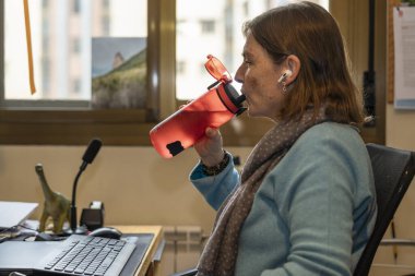 Yeniden kullanılabilir bir şişeden su içen olgun bir kadın ev ofisinde rahat bir şekilde çalışırken, iş ve refahı dengeliyor.