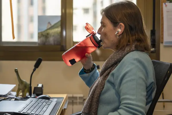 Yeniden kullanılabilir bir şişeden su içen olgun bir kadın ev ofisinde rahat bir şekilde çalışırken, iş ve refahı dengeliyor.