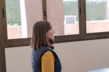 Sınıfta dikilirken pencereden dışarı bakan kadın öğretmen, dersleri yansıtıyor ve öğrenme ortamı ile iç içe.