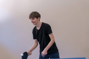 Genç, parlak bir odada masa tenisi oynuyor, topa ustaca kürekle vuruyor, konsantrasyon ve hareket gösteriyor.