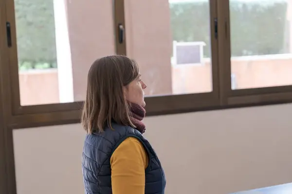 Sınıfta dikilirken pencereden dışarı bakan kadın öğretmen, dersleri yansıtıyor ve öğrenme ortamı ile iç içe.