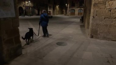 Kretas 'ın ortaçağ kaldırım taşlı sokaklarında tasmalı siyah köpeği gezdiren kadın, sakin İspanyol köyündeki yumuşak gölgeler ve mimari ışıklarla aydınlatılıyor.