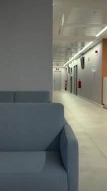 Modern bir hastane ya da klinik binasında uzun ve temiz bir koridorun yanında rahat bir kanepesi olan boş bir bekleme odası. Kimse yok.