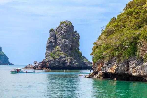 Mu Ko Ang Tangası, Tayland, turkuaz sudan yükselen yüksek bir kaya oluşumuyla manzaralı kıyı manzarası. Bir tekne yüzen bir rıhtıma yanaşmış, üzerinde insanlar yürüyor. Yeşil tepeler ve uçurumlar manzarayı çerçeveler, gökyüzü açık yumuşak mavi tonlarla.