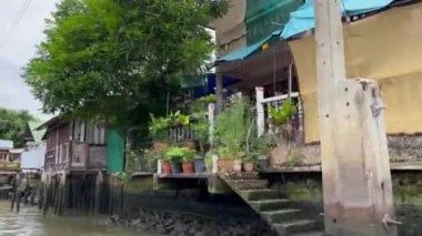 Tekne ve nehirden görüldüğü gibi nehir kıyısı boyunca sıralı evler. Evlerde, toplumun eşsiz karakterini yansıtan renk ve dokuların karışımı yer alıyor. Yeşillik ahşap yapılardan sarkıyor. Bangkok kanallarında çekildi.