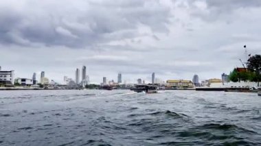 Görüntü, bulutlu bir gökdelenin altında yükselen gökdelenlere karşı teknelerle hareketli bir su yolu yakalıyor. Sahne, dinamik şehir yaşamını ve nehir kıyısının manzaralı güzelliğini yansıtıyor. Chao Phraya Nehri, Bangkok, Tayland.