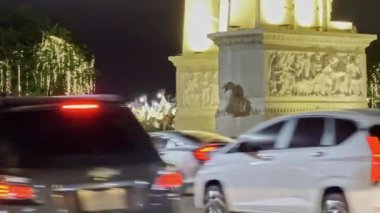 Ağaçlardaki dekoratif ışıklarla çevrili bir heykelle süslenmiş güzel aydınlatılmış bir gece manzarası. Ortam geçen araçlarla gelişiyor, dinamik bir şehir atmosferi yaratıyor..