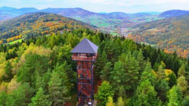 Parlak sonbahar ağaçlarıyla çevrili bir gözetleme kulesinin göz kamaştırıcı hava manzarası. Manzara, sonbahar mevsiminde doğanın güzelliğini gözler önüne seren yuvarlanan tepeler ve renkli bir desen içeriyor. Drone, hava aracı..