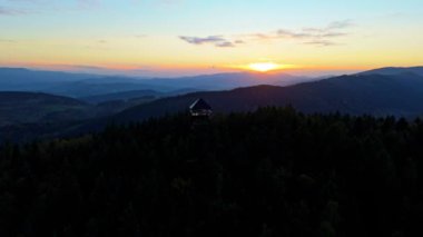 Arkasında güneşin battığı yemyeşil ormanlarla çevrili bir dağın tepesinden çarpıcı bir hava manzarası. Gün ışığı engebeli dağ manzarasında alacakaranlığa yol açarken gökyüzünün renkleri güzel bir şekilde değişiyor. Uçan İHA.