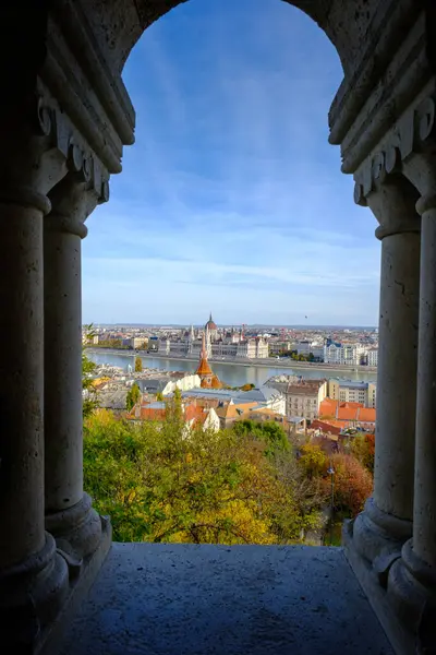 Bu pitoresk sahne, Tuna Nehri boyunca uzanan canlı Budapeşte şehrini gösteren bir kale kemerinden çarpıcı bir görüntü yakalıyor. Tarihi mimari ve doğal güzelliğin karışımı büyüleyici bir manzara yaratır..