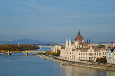 Tuna Nehri kıyıları boyunca uzanan Macar Parlamentosu Binası, açık mavi gökyüzü altında çarpıcı mimarisini sergiliyor. Köprü ve nehir Budapeşte 'nin güzel manzarasını güzelleştiriyor..