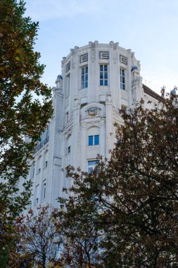 Bu resim, Budapeşte 'de verimli ağaçlar tarafından çerçevelenmiş bir Art Deco binasının karmaşık mimari özelliklerini yakalar. Zarif tasarım, modernlik ve geleneğin bir karışımını sergileyerek kentin zengin kültürel mirasını yansıtıyor..