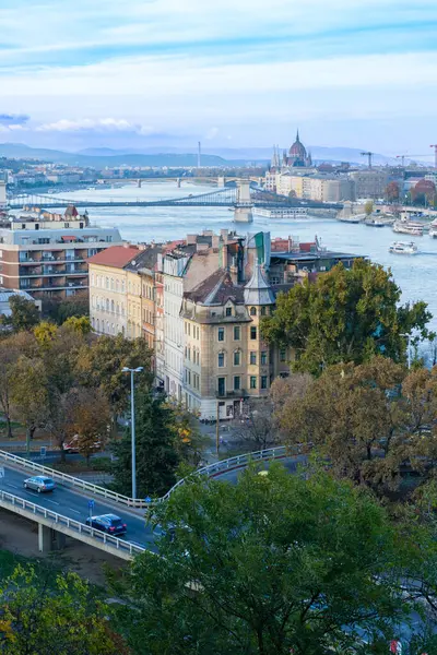 Tuna Nehri Budapeşte 'deki zarif binaların önünden akıyor. Ön planda ağaçlar ve yollar yer alırken, ufuk çizgisi tarihi mimari ve çağdaş gelişime dair bir ipucu yer alıyor..