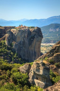 Görkemli kaya oluşumları ve yemyeşil yeşilliklerle çevrili yüksek bir uçuruma tünemiş bir manastırın nefes kesici manzarası. Arkaplanda açık mavi gökyüzünün altındaki uzak dağlar yer alıyor. Meteora 'nın çarpıcı manzarasını gözler önüne seriyor..